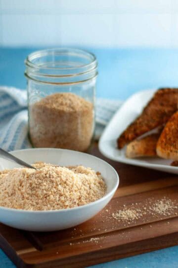 Bowl of sprinkle parmesan next to a plate of garlic bread sitting on a wooden cutting board.