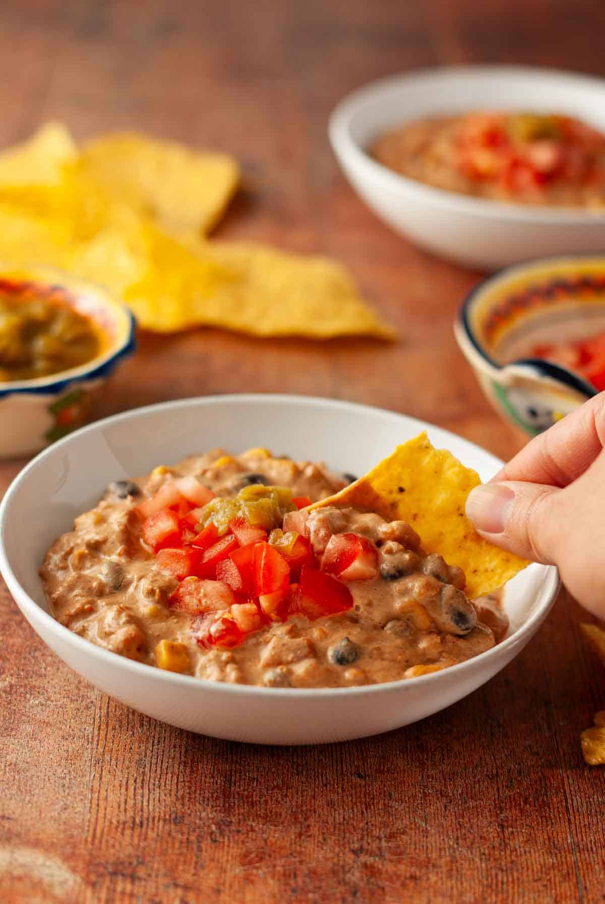 A hand dipping a tortilla chip into a bowl of southwest vegan chili on a wooden table.