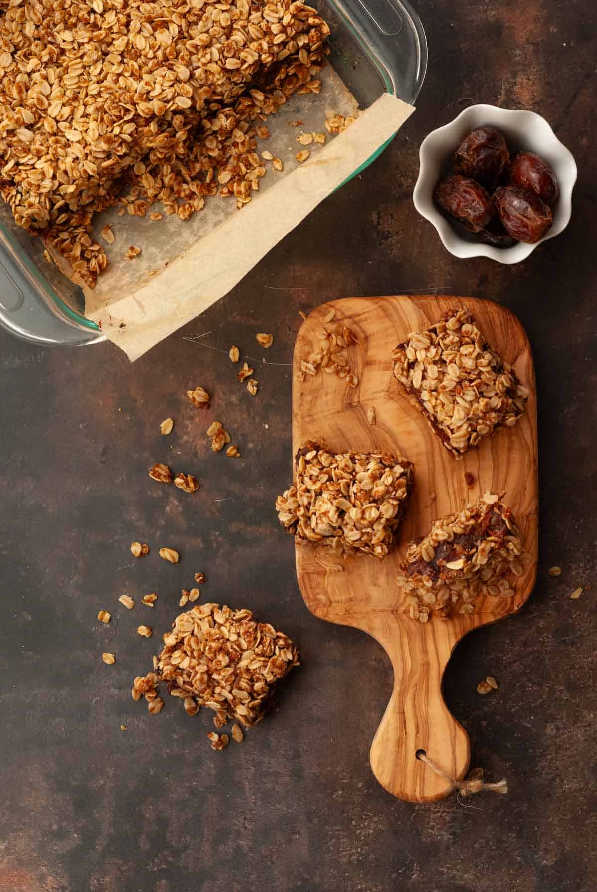 An overhead view of baked vegan date bars in a baking dish and some cut bars shown on a wooden board with a decorative bowl of dates.