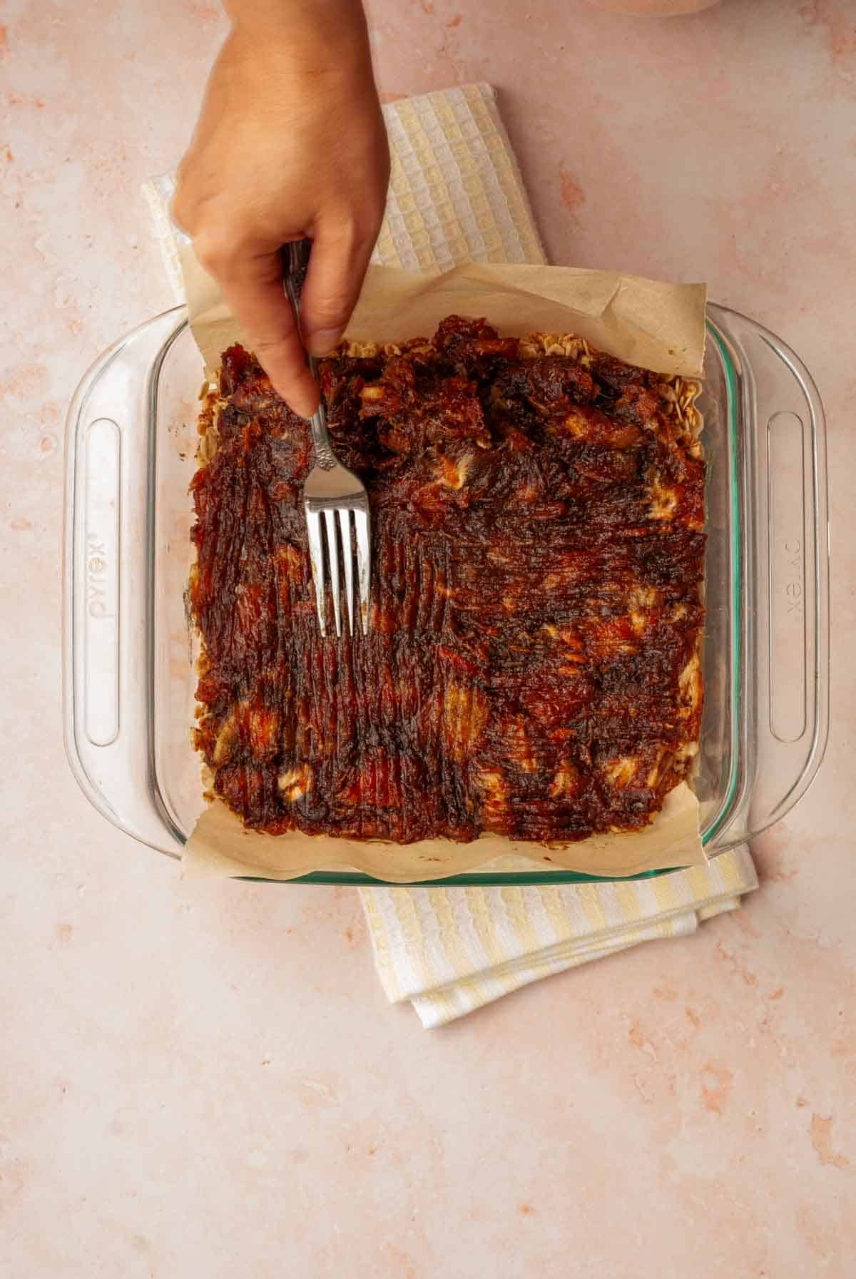 A hand shown spreading softened medjool dates with a fork in a baking dish.