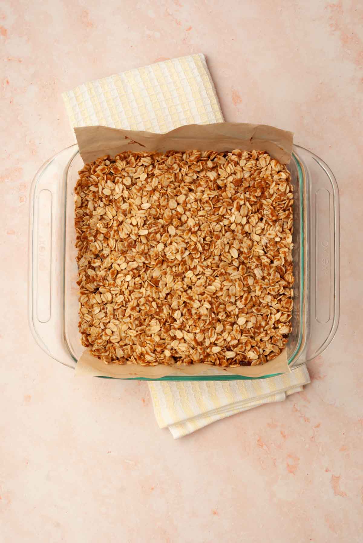 Fully baked vegan date bars in a baking dish lined with parchment sitting on a white and yellow striped tea towel.