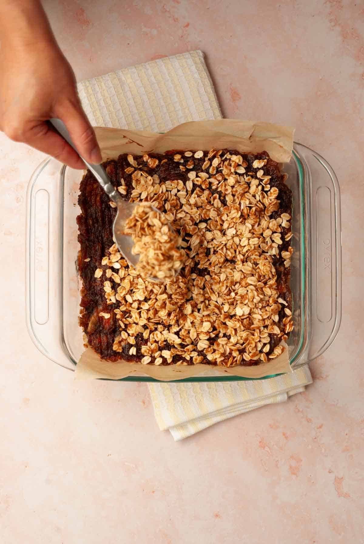 A hand shown sprinkling oat and coconut mixture over date bars in a baking dish.