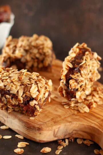 Three vegan date bars covered in chewy oats on a wooden board with a bowl of dates in the background.