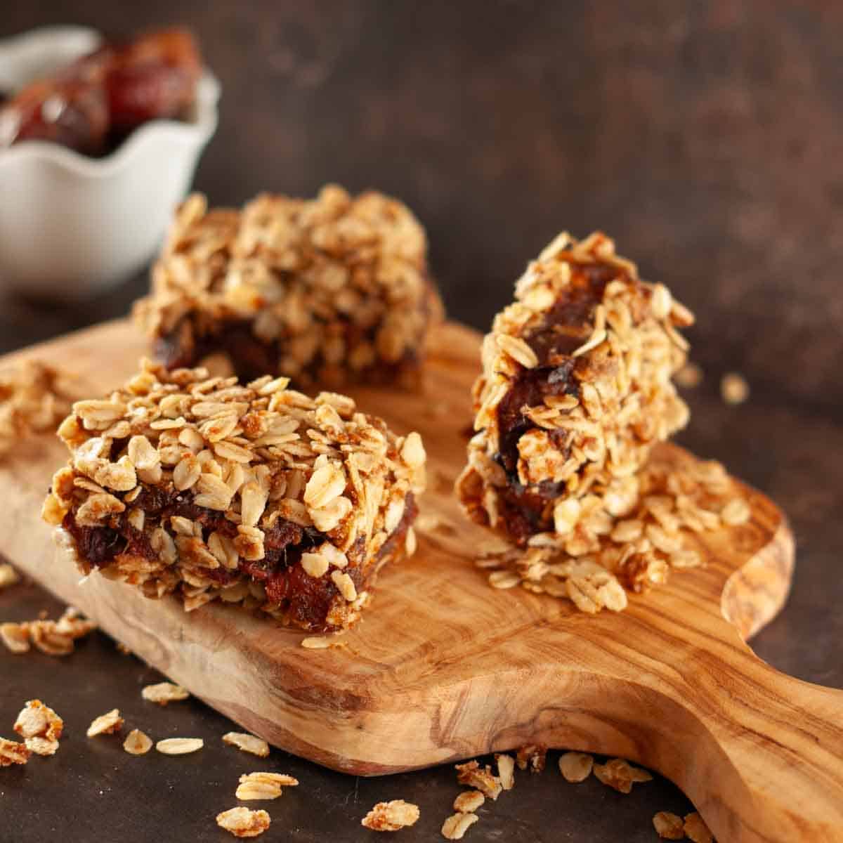 Three vegan date bars covered in chewy oats on a wooden board with a bowl of dates in the background.