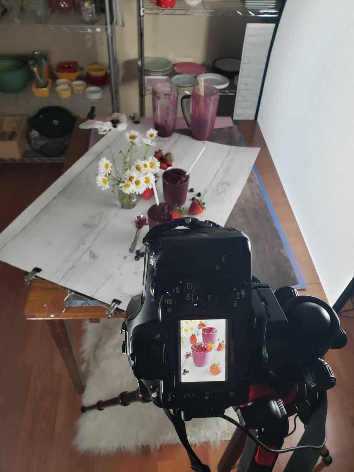 The smoothies set up on a backdrop with daisies and fresh strawberries and blueberries with the DSLR camera showing the recently captured image.