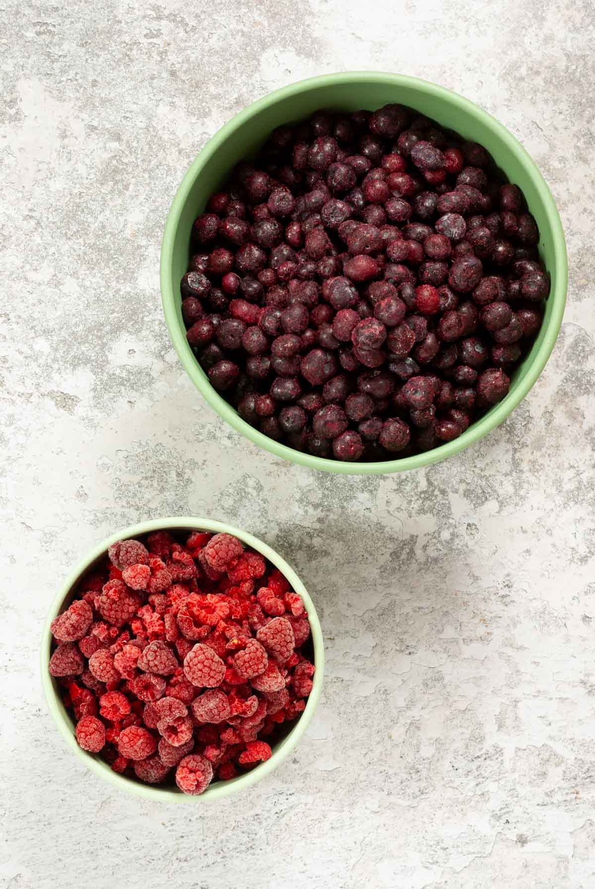 A large bowl of frozen Blueberries and a smaller bowl of frozen Raspberries.