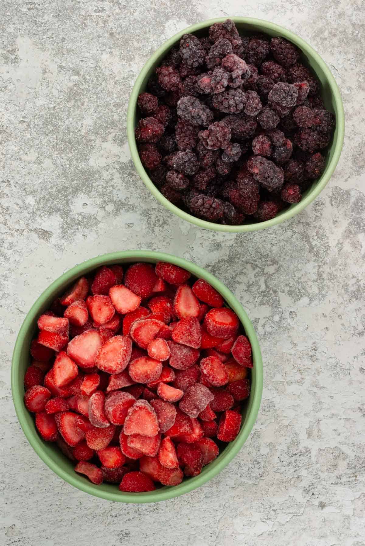 A large bowl of frozen strawberries and a smaller bowl of frozen blackberries.