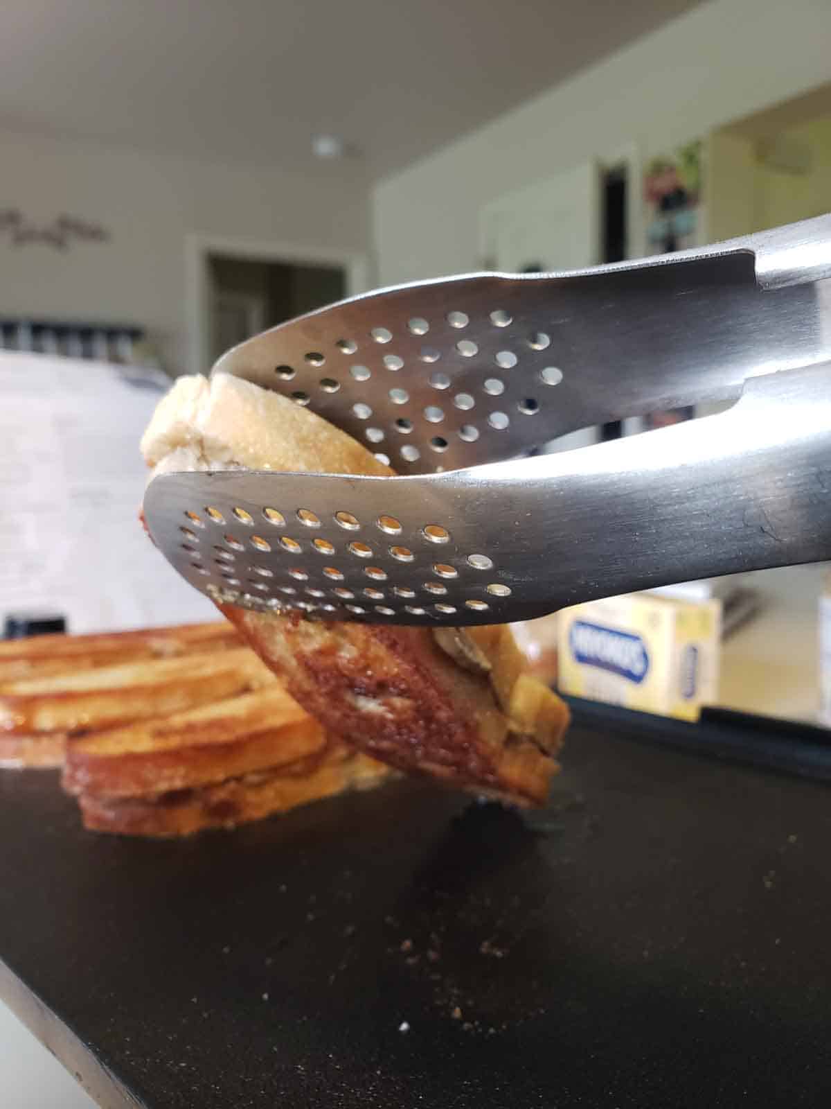 A pair of tongs showing a sandwich being flipped on a griddle.