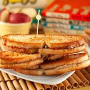 3 Grilled sunbutter & Jelly sandwiches stacked on a white plate on a bamboo placemat with children's books in the background.