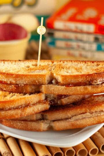 3 Grilled sunbutter & Jelly sandwiches stacked on a white plate on a bamboo placemat with children's books in the background.