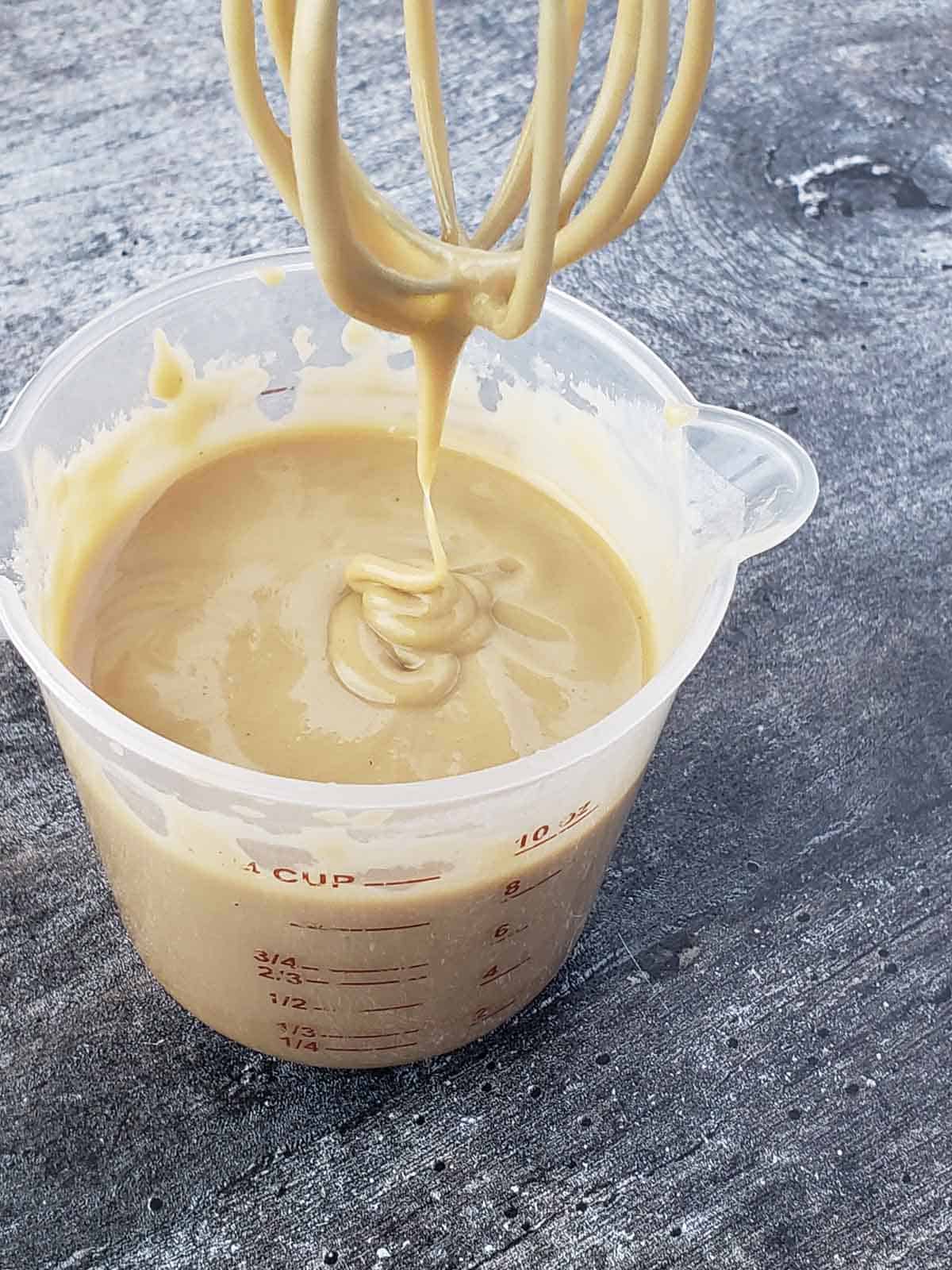 A whisk being pulled out of a cup of lemon tahini sauce to show thickness and texture.
