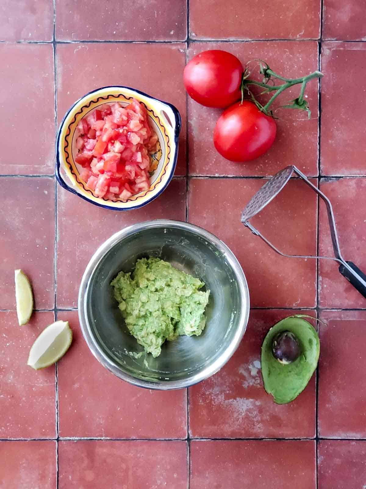 A bowl of chopped tomatoes and a bowl of mashed avocado with limes, whole tomatoes, and a potato masher next to them.