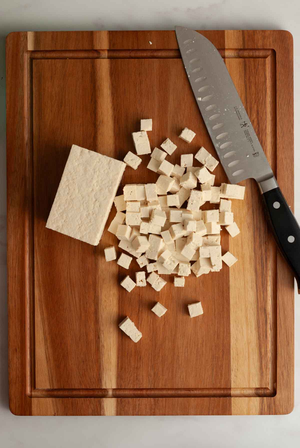 A pile of chopped tofu on a wooden cutting board with a large chopping knife.