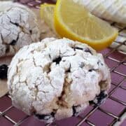 A lemon blueberry crinkle cookie on a cooling rack showing details of the powdered sugar cracking. Lemon slices and more cookies in the background.