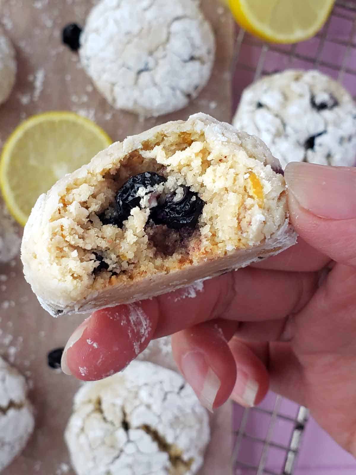 A hand holding a lemon blueberry crinkle cookie with a bite taken out of it. More cookies and lemon slices in the background.
