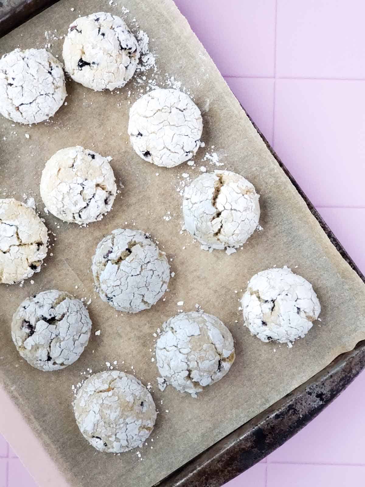 Freshly baked lemon blueberry crinkle cookies on a parchment lined baking tray on a pink tile flat lay.