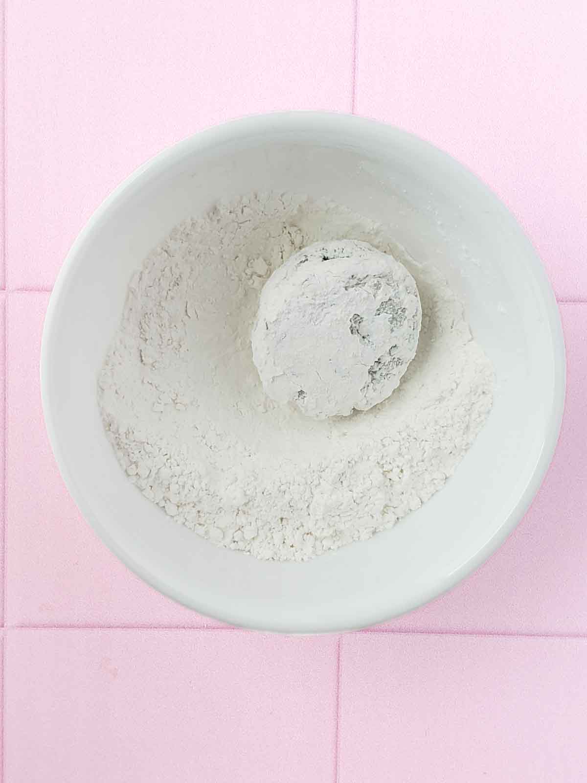 A bowl of powdered sugar and a cookie scoop shown rolling in the sugar to evenly coat it.