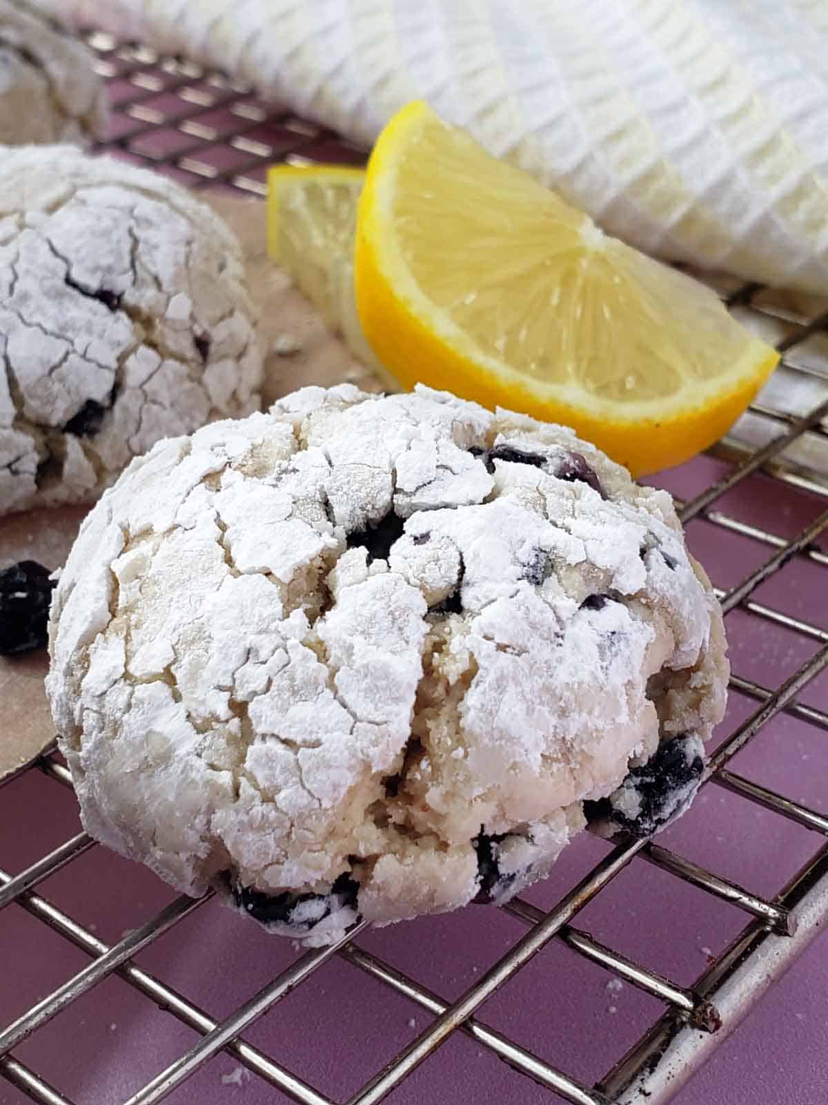 A lemon blueberry crinkle cookie on a cooling rack showing details of the powdered sugar cracking. Lemon slices and more cookies in the background.