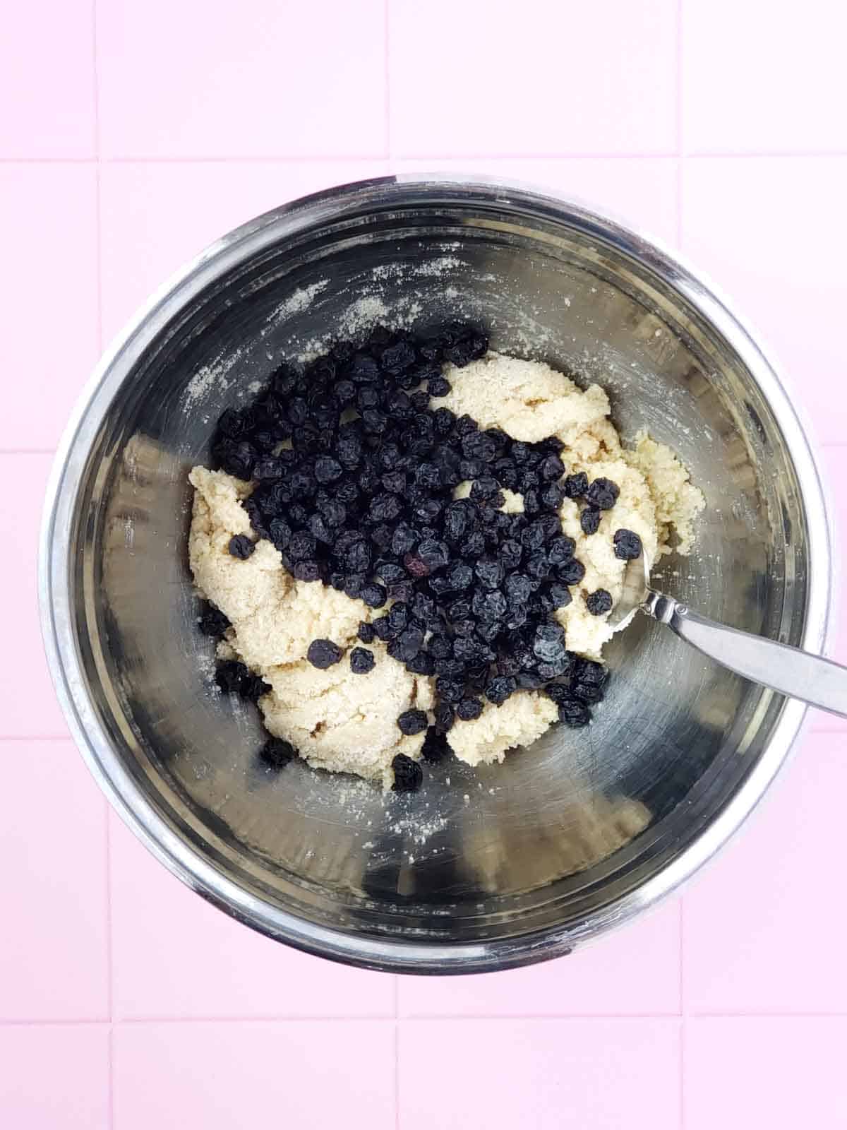 Blueberries shown being mixed into the cookie dough in a mixing bowl with a spoon.
