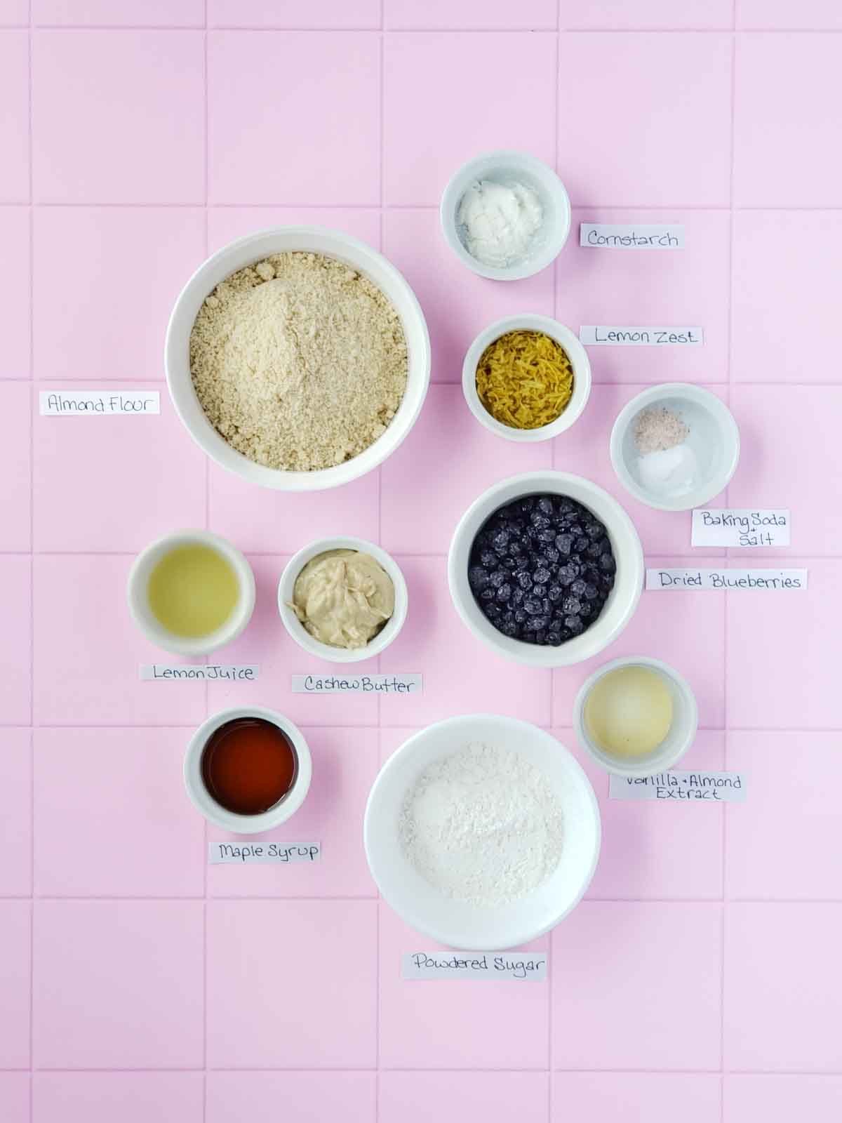 All lemon blueberry crinkle cookie recipe ingredients shown in separate bowls with labels on a pink tile background.