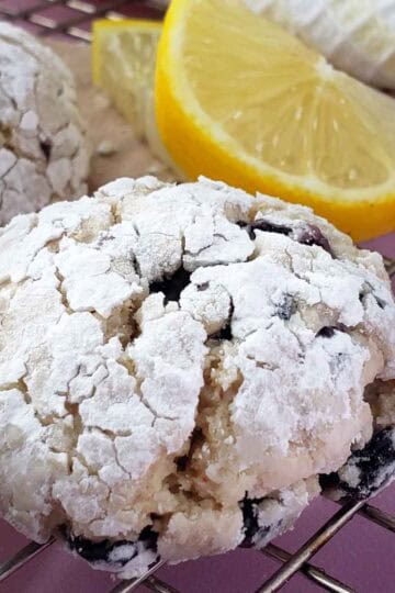 A lemon blueberry crinkle cookie on a cooling rack showing details of the powdered sugar cracking. Lemon slices and more cookies in the background.
