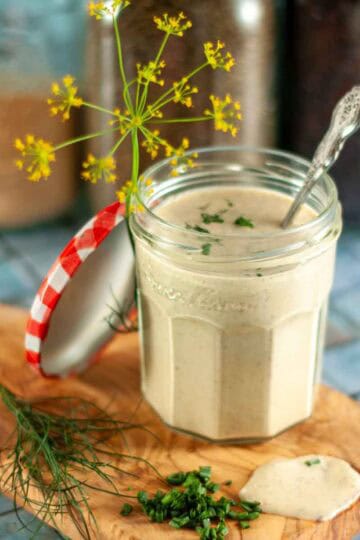 A jar of creamy ranch dressing on a wooden board with fresh dill and chives and a dill flower, with spices in the background.