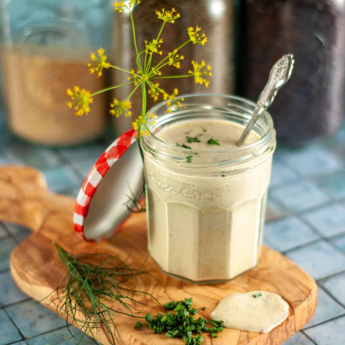 A jar of creamy ranch dressing on a wooden board with fresh dill and chives and a dill flower, with spices in the background.