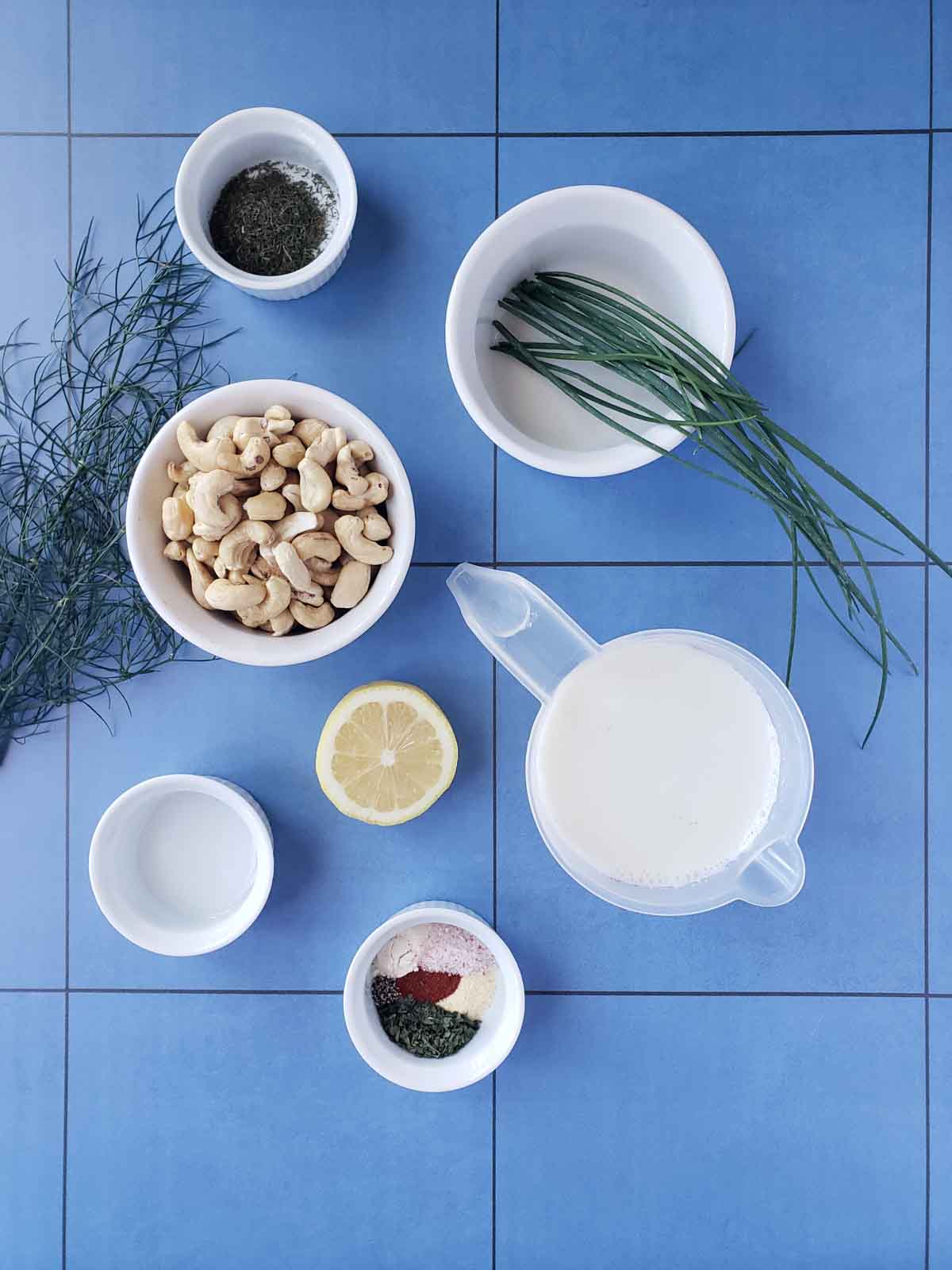 Dairy free ranch dressing ingredients including raw cashews, fresh herbs, plant milk, fresh lemon, and vinegar on a blue tile flat lay.