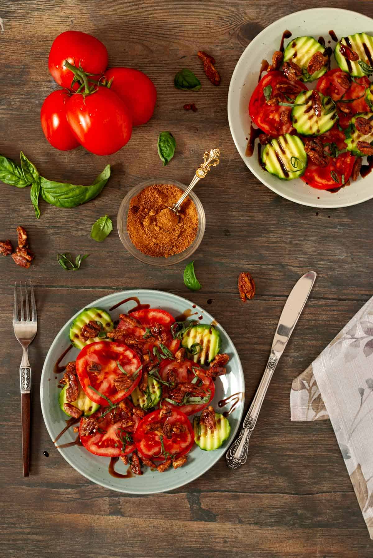 Table top view of tomato, basil, cucumber salad on two plates with basil leaves and vine tomatoes on table.