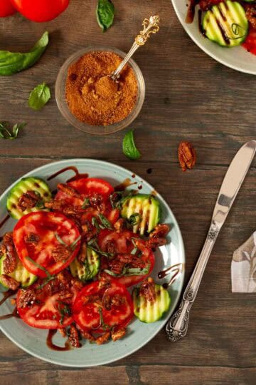 Table top view of tomato, basil, cucumber salad on two plates with basil leaves and vine tomatoes on table.