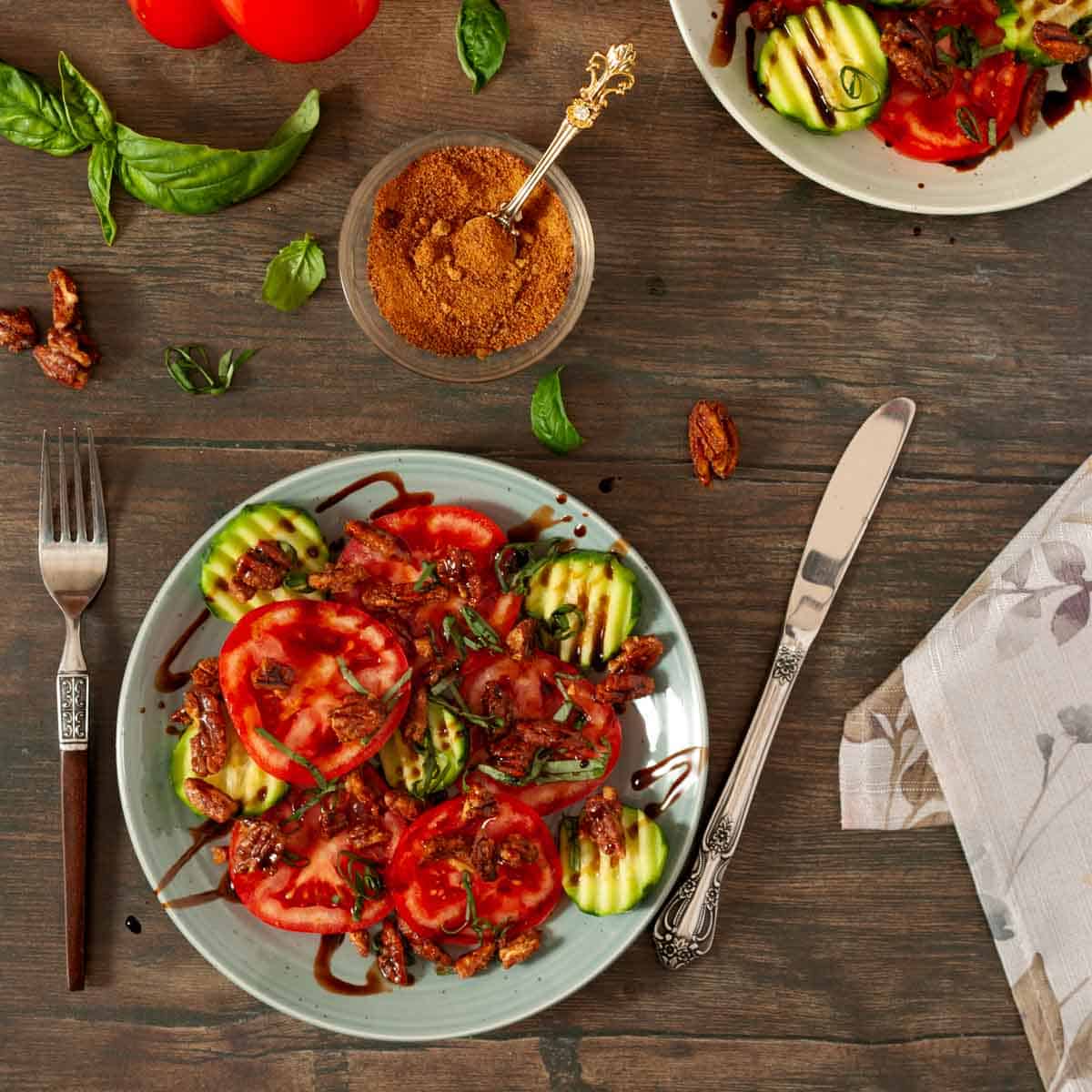 Table top view of tomato, basil, cucumber salad on two plates with basil leaves and vine tomatoes on table.