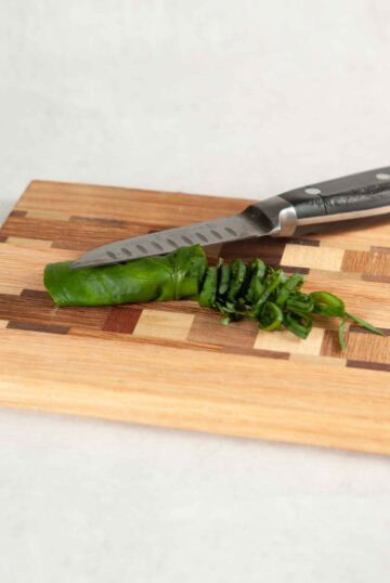 Rolled basil leaves on a wooden cutting board with a knife demonstrating how to julienne.