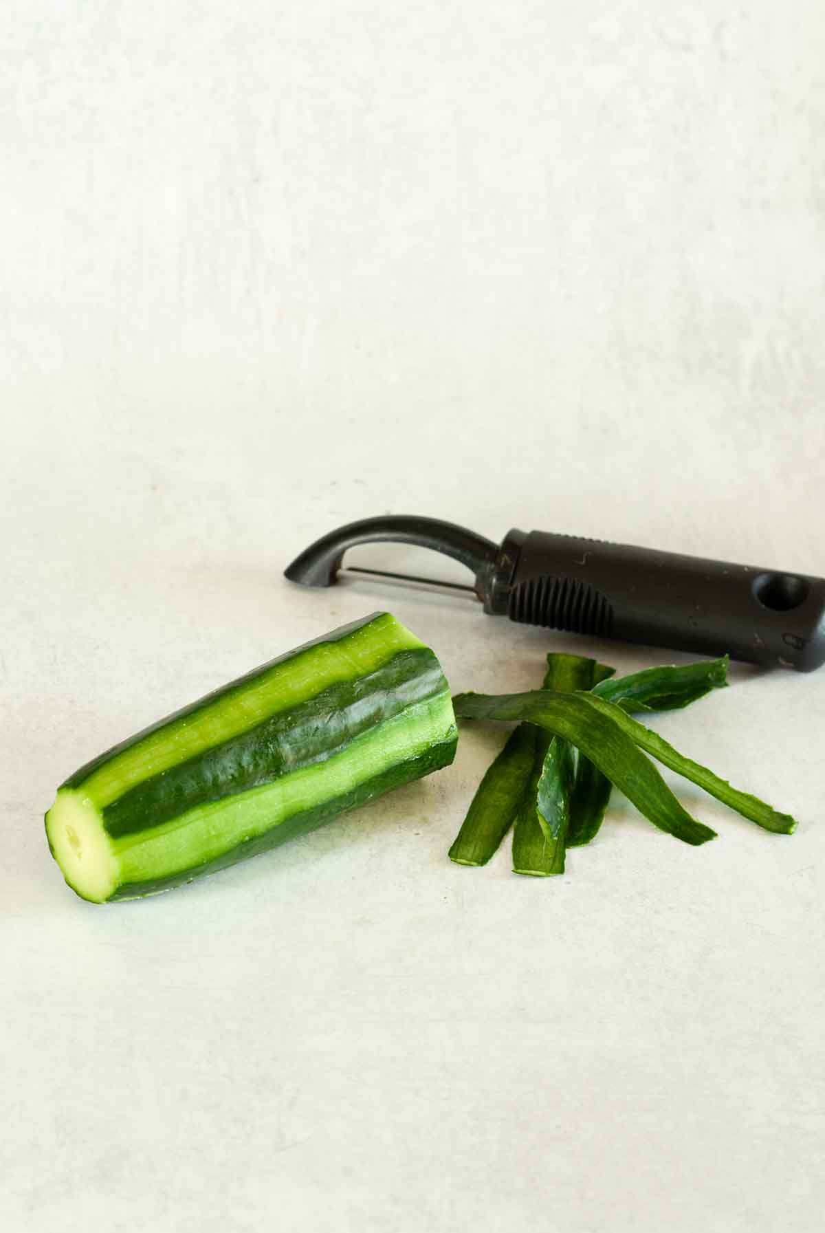 A cucumber partially peeled, cucumber peelings, and a vegetable peeler.