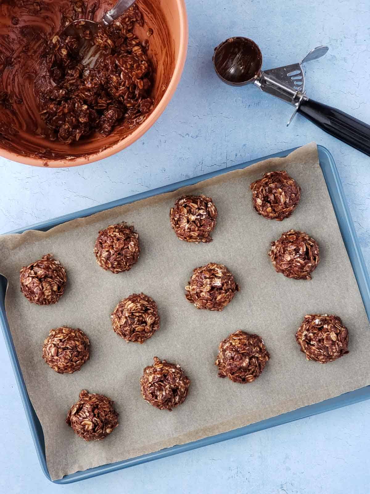 A mixing bowl with cookie batter and a used cookie scoop shown next to a tray filled with the scooped balls of batter before being pressed.