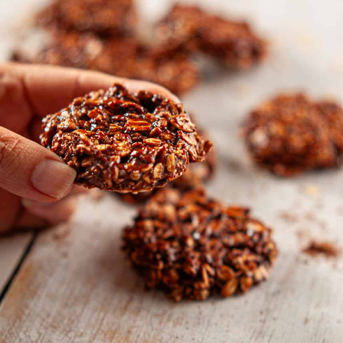 A hand holding a single chocolatey no bake refrigerator cookie with more cookies in the background on a wooden table.