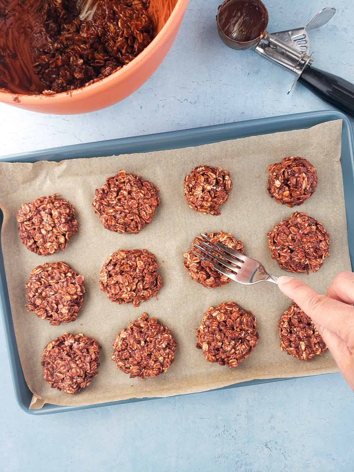 A hand shown using a fork to press each ball of batter into a flatter cookie shape before refrigerating.