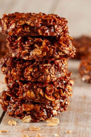 A stack of 5 chewy chocolate refrigerator cookies on a wooden table with some raw oats scattered about.