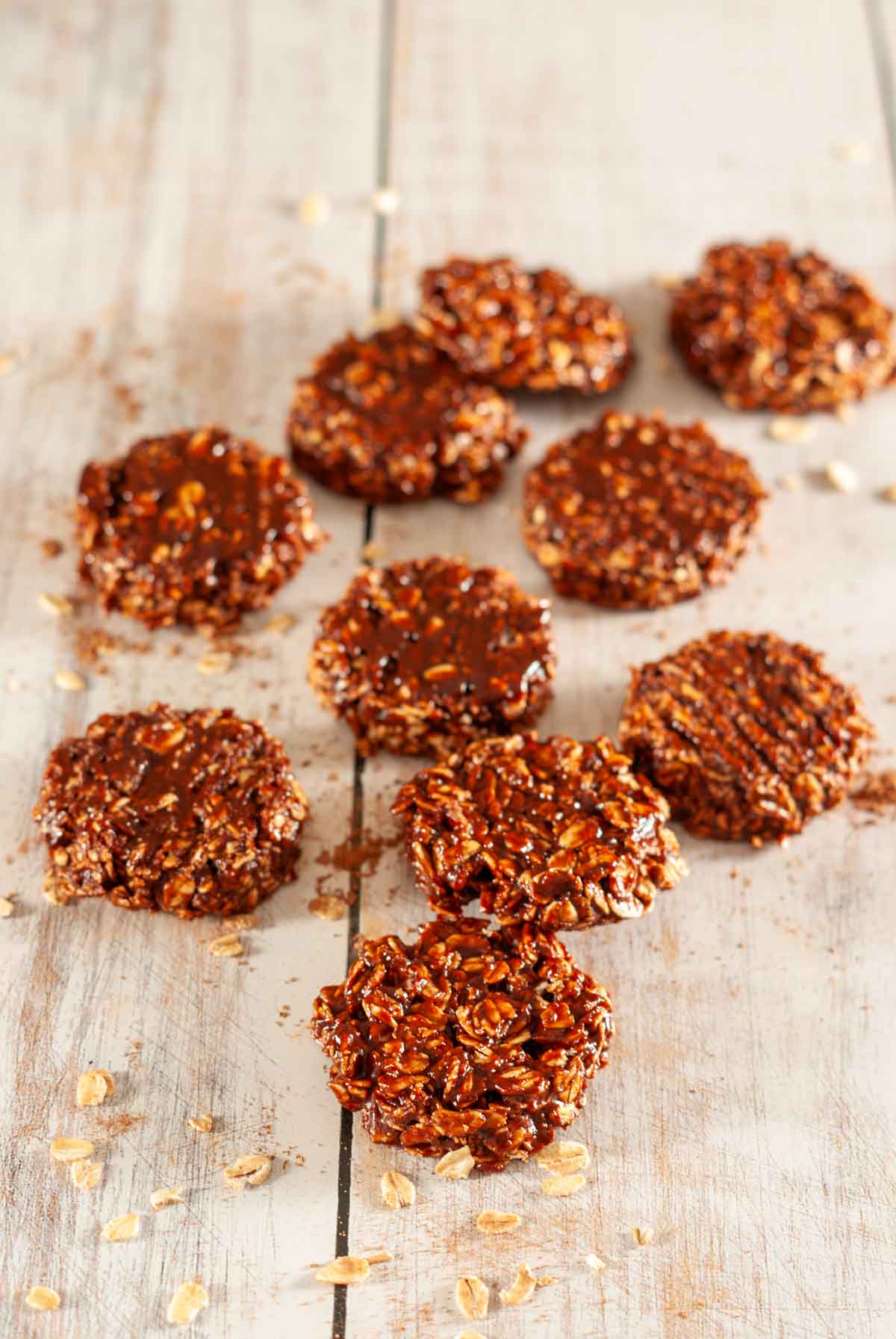 Ten chocolate cookies laid out on a wooden table with some raw oats and cocoa powder.