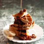 Vegan banana buckwheat waffles stacked with pecans, banana slices, and maple syrup dripping down.