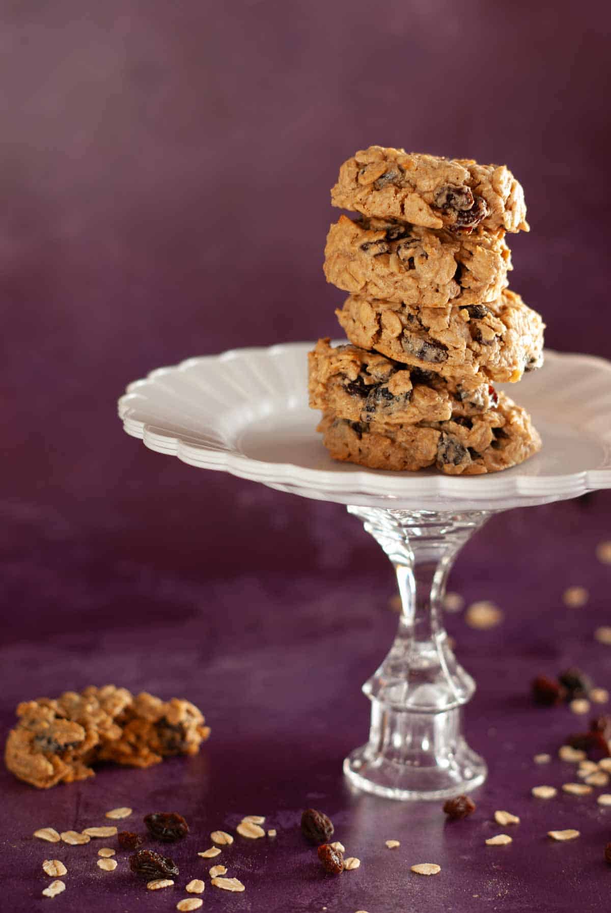 Stack of oatmeal raisin cookies on white pedestal with raisins scattered.