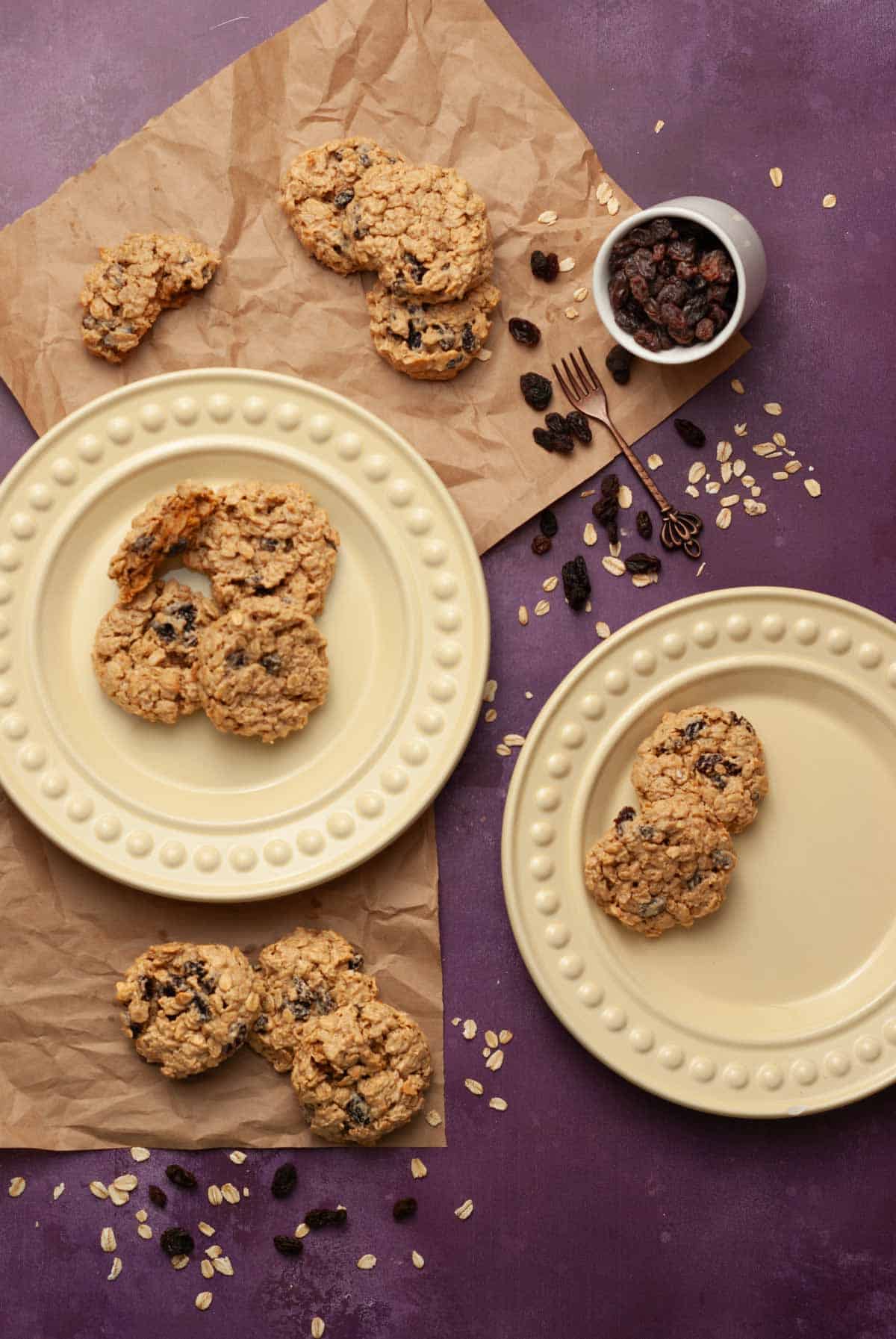 Homemade oatmeal raisin cookies on plates with oats and raisins scattered on a purple background.