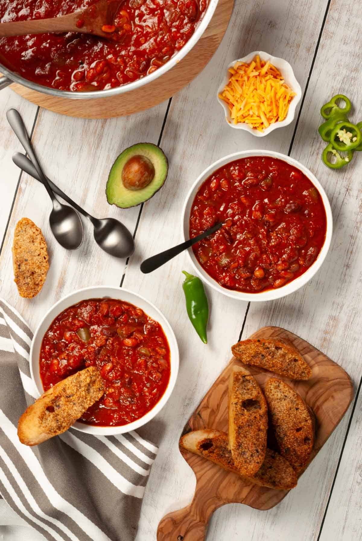 Overhead view of vegan chili in bowls and pot with bread, avocado, and toppings on a white rustic table.