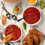 Overhead view of vegan chili in bowls and pot with bread, avocado, and toppings on a white rustic table.