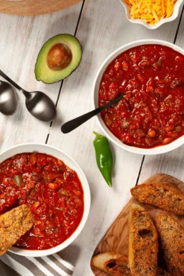 Overhead view of vegan chili in bowls and pot with bread, avocado, and toppings on a white rustic table.