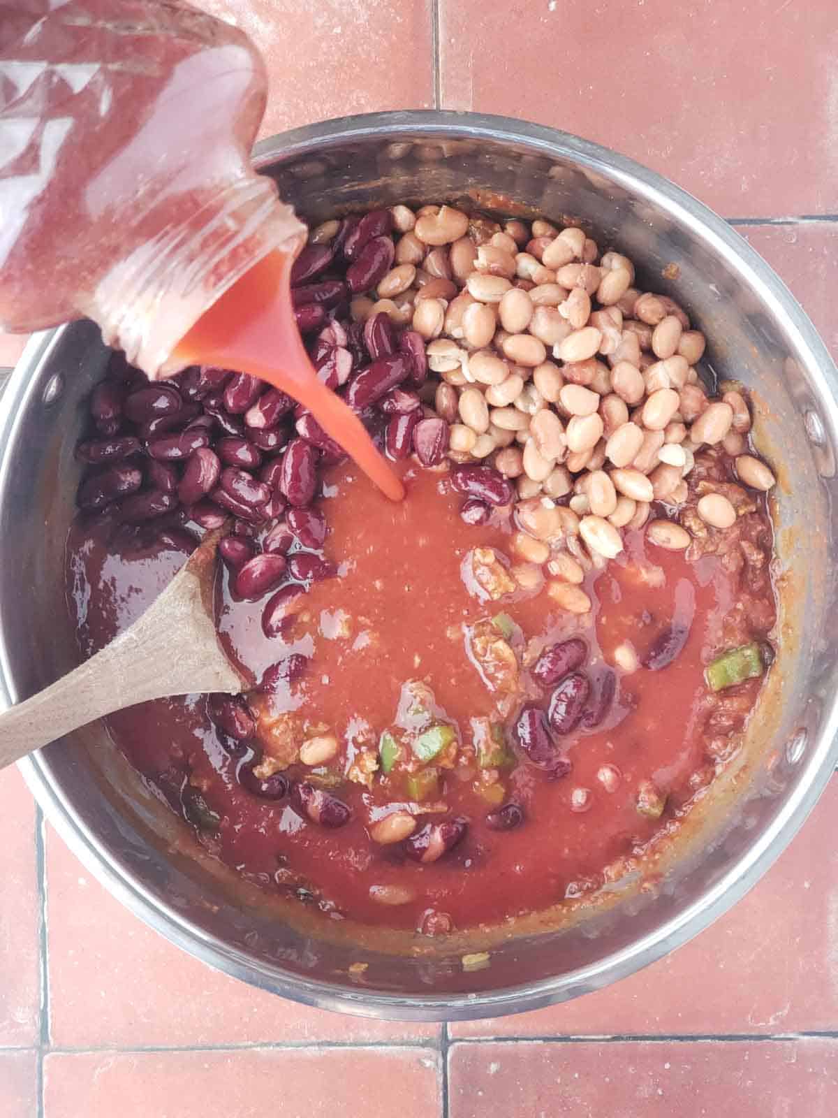 Vegetable juice being poured into pot with beans and tomatoes, vegan meat, and veggies.