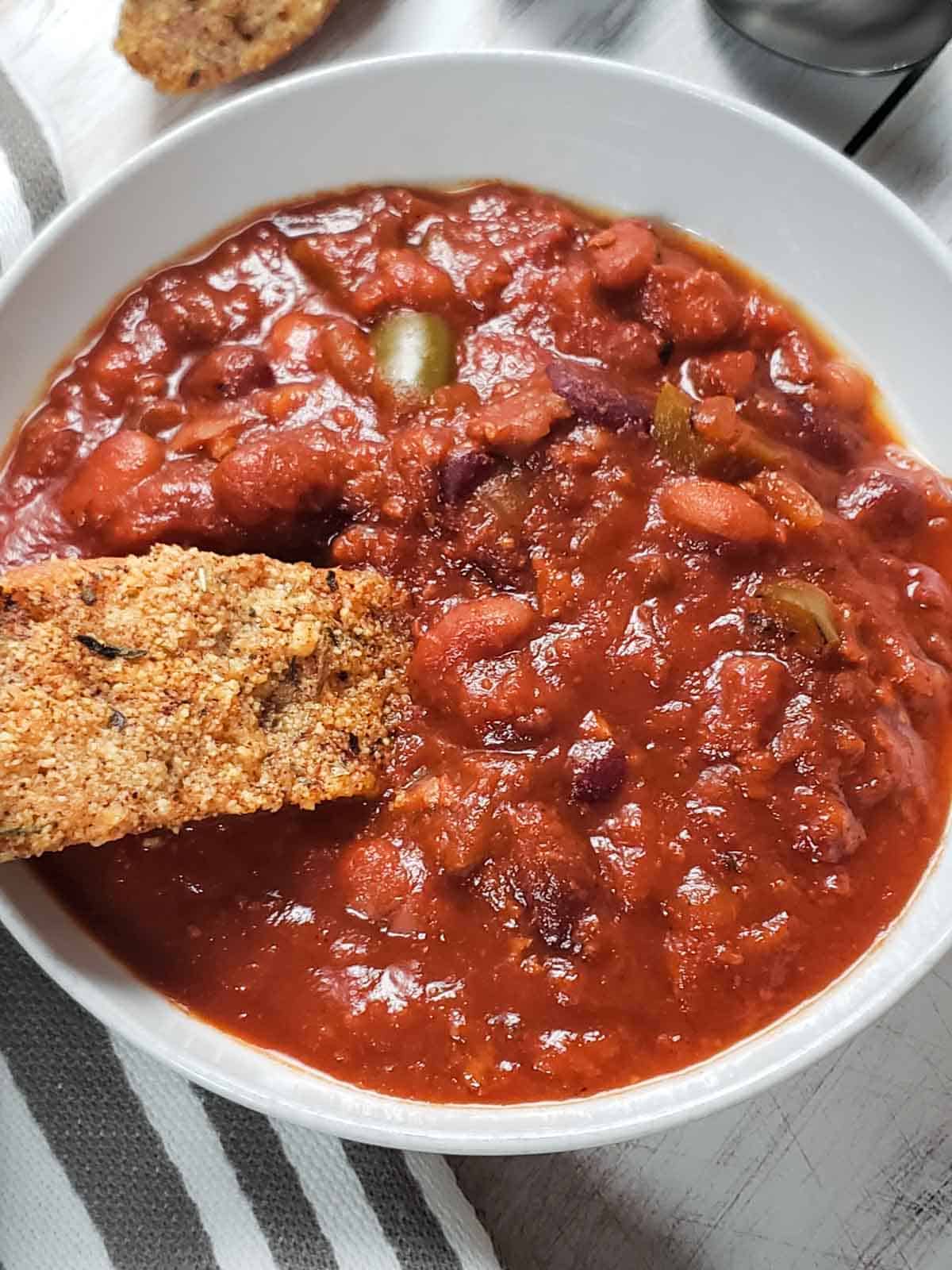 Close-up of thick vegan chili in white bowl with slice of toasted garlic bread for dipping.