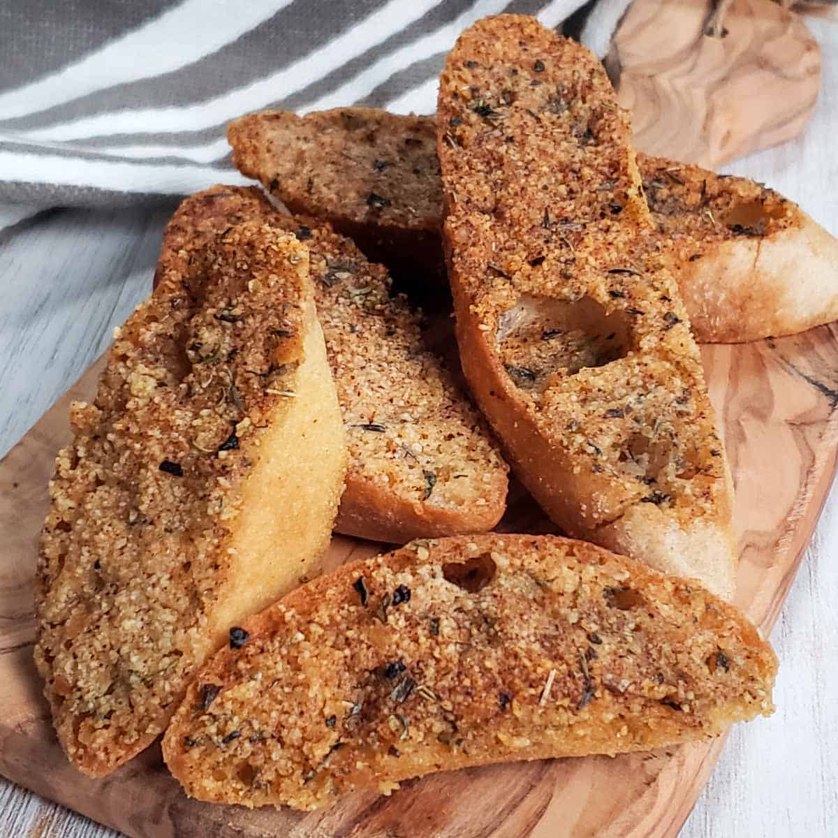 Several garlic toast slices served on a wooden board, crispy and golden brown with gray and white striped towel in the background.