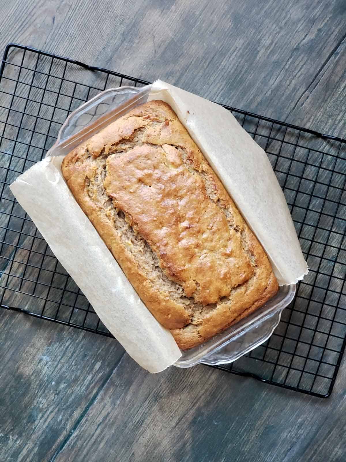 Freshly baked loaf of golden banana bread cooling in a parchment-lined glass loaf pan on a wire rack.