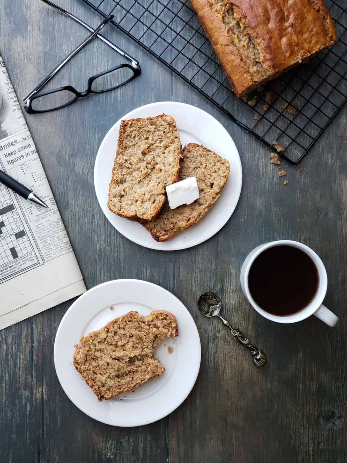 Overhead view of sliced banana bread on white plates with coffee and a newspaper on a rustic wooden table.