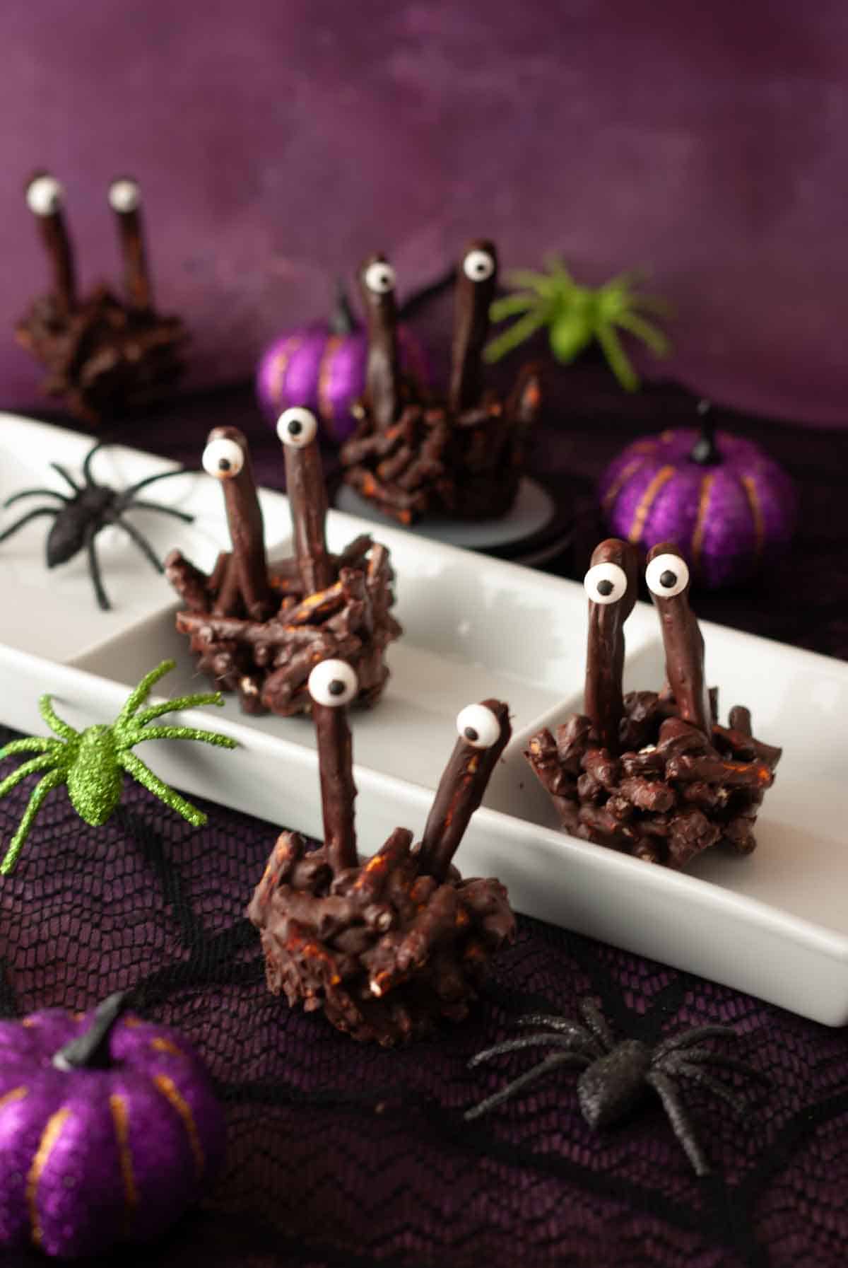 Chocolate-covered pretzel monster clusters on white tray with glittery Halloween spiders and pumpkins.